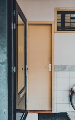A narrow entryway featuring a closed beige door with silver hardware. The area to the left includes a glass window, reflecting part of the scene. On the right, a section of white tiles with a gray border is visible, preceded by a partial view of a bicycle wheel. Above the door, there is a small window with horizontal panes.