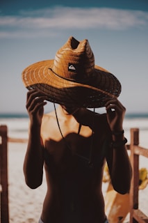 A person standing shirtless near the beach with a large woven straw hat obscuring their face. The background shows a sandy beach and blue sky, with part of a wooden fence visible. The lighting suggests a warm, sunny day, casting shadows on the person’s body and the hat.