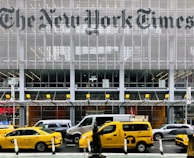 Yellow taxis are parked and driving in front of a modern building with large lettering displaying a well-known newspaper name. The facade is made up of metal slats and glass, creating a sleek and urban atmosphere. Pedestrians and various vehicles are also present in the busy city environment.