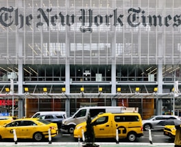 Yellow taxis are parked and driving in front of a modern building with large lettering displaying a well-known newspaper name. The facade is made up of metal slats and glass, creating a sleek and urban atmosphere. Pedestrians and various vehicles are also present in the busy city environment.