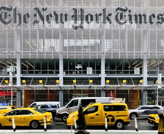 Yellow taxis are parked and driving in front of a modern building with large lettering displaying a well-known newspaper name. The facade is made up of metal slats and glass, creating a sleek and urban atmosphere. Pedestrians and various vehicles are also present in the busy city environment.