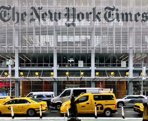 Yellow taxis are parked and driving in front of a modern building with large lettering displaying a well-known newspaper name. The facade is made up of metal slats and glass, creating a sleek and urban atmosphere. Pedestrians and various vehicles are also present in the busy city environment.