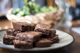 A plate stacked with chocolate brownies is placed on a wooden table. The brownies are richly textured with a slightly crumbly top, attracting attention in the soft focus background.