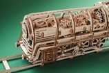 A detailed wooden model of a locomotive displaying intricate gears and mechanical components. The model is crafted using light-colored wood with visible grain patterns and is positioned on a small wooden track. The background is a solid green color, highlighting the craftsmanship of the model.