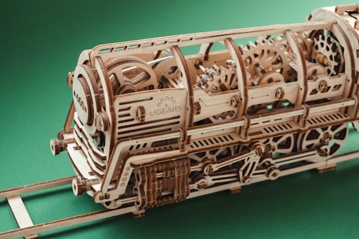 A detailed wooden model of a locomotive displaying intricate gears and mechanical components. The model is crafted using light-colored wood with visible grain patterns and is positioned on a small wooden track. The background is a solid green color, highlighting the craftsmanship of the model.