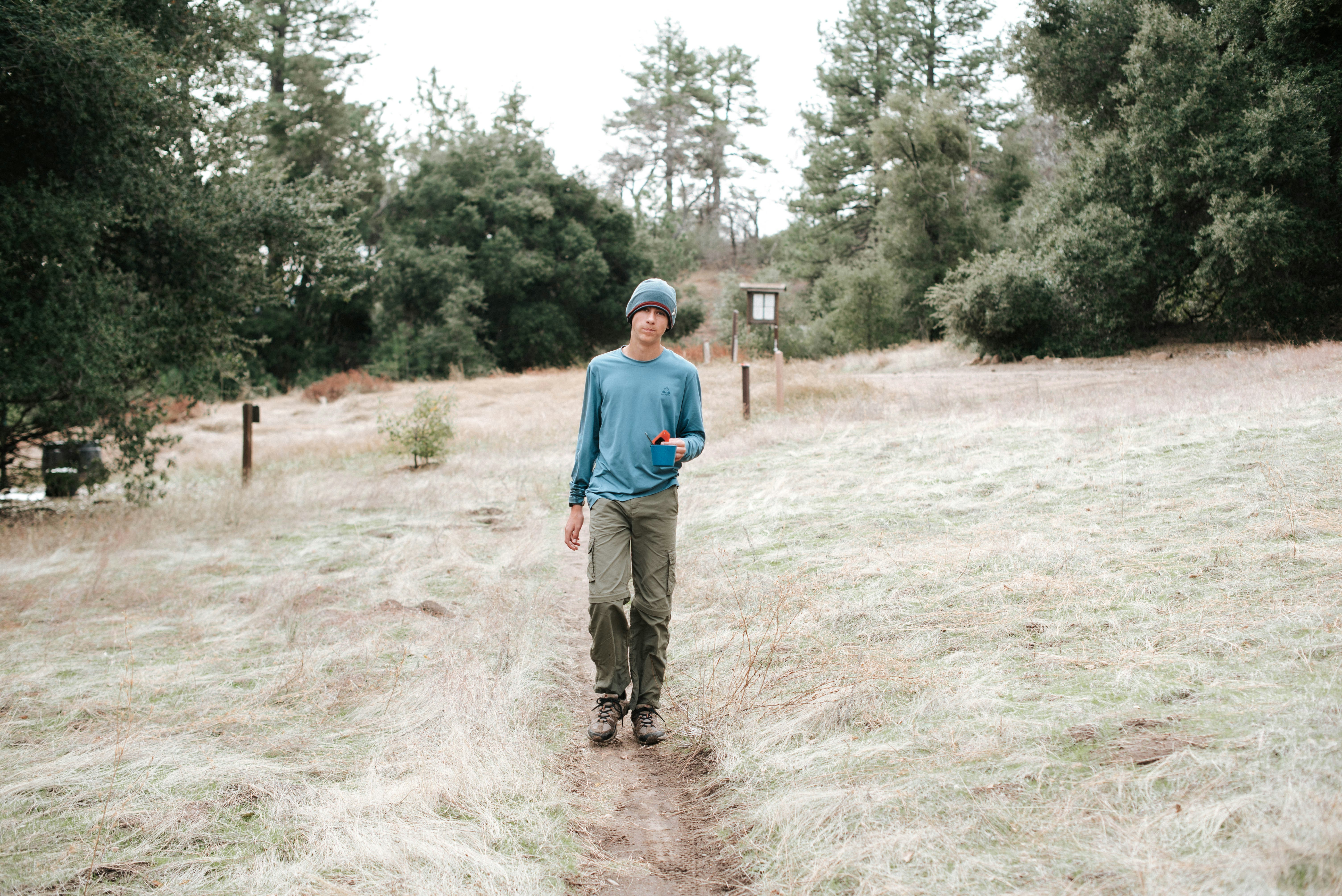 A hiker in a blue long-sleeve top, green trousers and a knit cap walks toward the camera on a dirt path through pale dry grass, pines and oaks behind