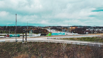 A highway interchange with several semi-trucks transporting shipping containers. The environment is overcast with visible clouds and a distant urban landscape. Grass and sparse trees line the foreground, and various roads intersect and curve.