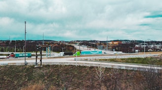 A highway interchange with several semi-trucks transporting shipping containers. The environment is overcast with visible clouds and a distant urban landscape. Grass and sparse trees line the foreground, and various roads intersect and curve.