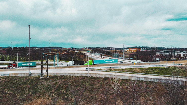 A highway interchange with several semi-trucks transporting shipping containers. The environment is overcast with visible clouds and a distant urban landscape. Grass and sparse trees line the foreground, and various roads intersect and curve.