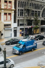A blue news van labeled 'Eyewitness News' is parked on a city street. The background features old, ornate buildings with large windows and balconies. Several cars are parked along the street with people standing on the sidewalk.