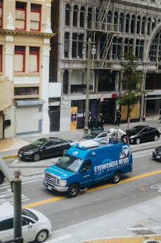 A blue news van labeled 'Eyewitness News' is parked on a city street. The background features old, ornate buildings with large windows and balconies. Several cars are parked along the street with people standing on the sidewalk.