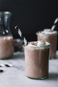 A glass jar filled with a creamy coffee-colored beverage topped with whipped cream and chocolate shavings. There is a black and white striped straw in the jar. In the background, another similar jar and a carafe of the same beverage are visible, all set against a dark backdrop.