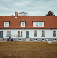 A long, single-story residential building with a red shingle roof and white brick walls. The facade features several evenly spaced windows with dark gray trim and two sets of steps leading to doorways. A bare tree stands in the foreground on a brown grass lawn, accompanied by a few objects such as a plastic toy car, a blue bin, and a planter.