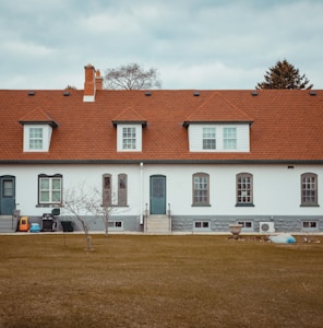 A long, single-story residential building with a red shingle roof and white brick walls. The facade features several evenly spaced windows with dark gray trim and two sets of steps leading to doorways. A bare tree stands in the foreground on a brown grass lawn, accompanied by a few objects such as a plastic toy car, a blue bin, and a planter.