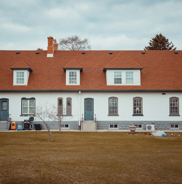 A long, single-story residential building with a red shingle roof and white brick walls. The facade features several evenly spaced windows with dark gray trim and two sets of steps leading to doorways. A bare tree stands in the foreground on a brown grass lawn, accompanied by a few objects such as a plastic toy car, a blue bin, and a planter.
