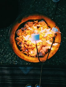 A warm campfire with glowing charcoals used for roasting marshmallows. Two marshmallows on skewers are being toasted over the flames, creating a cozy and inviting atmosphere. The ground beneath and a corner of a carpet is visible, suggesting an outdoor setting.