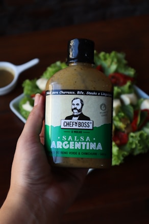 A hand is holding a bottle of salsa with a green and white label. The background includes a blurry image of a salad with greens and red elements, as well as a white sauce dish.