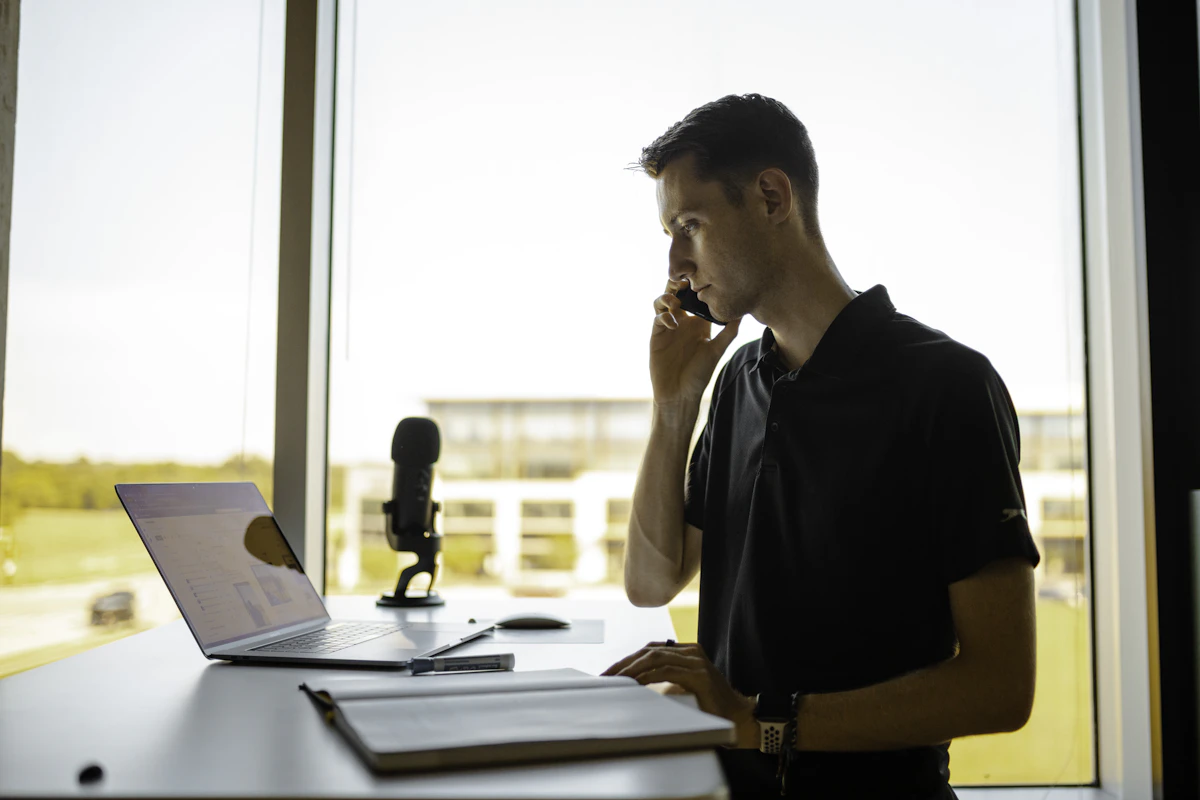 A man takes a phone call at his desk by a bright window, laptop open and a microphone beside it