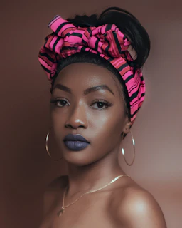 A woman with smooth skin wears a pink and black striped headscarf tied into a bow. She has bold blue lipstick and is accessorized with large hoop earrings and a delicate necklace. The background is a muted brown, complementing her appearance.