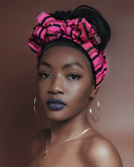 A woman with smooth skin wears a pink and black striped headscarf tied into a bow. She has bold blue lipstick and is accessorized with large hoop earrings and a delicate necklace. The background is a muted brown, complementing her appearance.