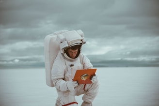An astronaut wearing a spacesuit is kneeling on a vast, flat expanse, resembling a salt flat or desert. The sky is overcast with clouds, adding a muted tone to the scene. The astronaut is holding a book or folder with a picture of an astronaut on it.