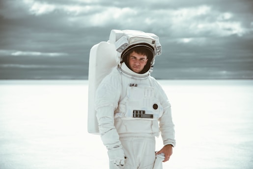 A person wearing a white astronaut suit stands against a vast, light-colored background with a cloudy sky above. The background is minimalist, emphasizing the futuristic and isolated setting.