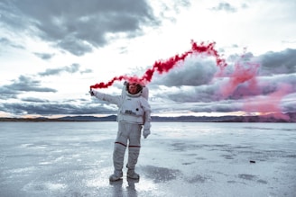 An astronaut stands on a vast salt flat, holding a red smoke flare in one hand. The sky is overcast with dramatic clouds, and the reflection of the scene is visible on the wet surface. The landscape is expansive and deserted, creating a surreal atmosphere.