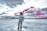An astronaut stands on a vast salt flat, holding a red smoke flare in one hand. The sky is overcast with dramatic clouds, and the reflection of the scene is visible on the wet surface. The landscape is expansive and deserted, creating a surreal atmosphere.