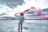 An astronaut stands on a vast salt flat, holding a red smoke flare in one hand. The sky is overcast with dramatic clouds, and the reflection of the scene is visible on the wet surface. The landscape is expansive and deserted, creating a surreal atmosphere.