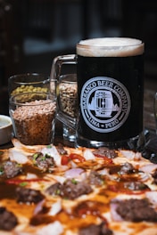 A large glass mug filled with dark beer is placed beside small glasses containing grains. In front of this setup is a pizza topped with various ingredients such as meat, onions, and herbs.