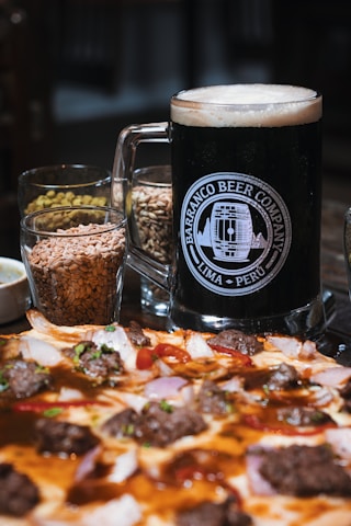 A large glass mug filled with dark beer is placed beside small glasses containing grains. In front of this setup is a pizza topped with various ingredients such as meat, onions, and herbs.