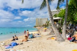 A vibrant beach scene with people sunbathing and swimming in the ocean. The foreground features sandy shores with towels and beach chairs scattered around. In the background, high-rise hotels line the coast, and tall palm trees sway gently in the breeze. The sky is partly cloudy with patches of bright blue.