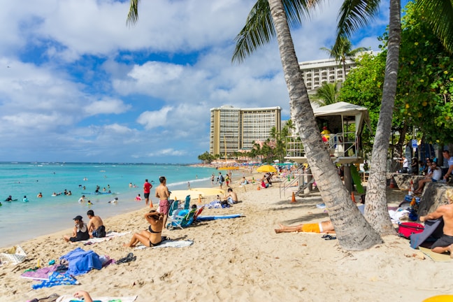 A vibrant beach scene with people sunbathing and swimming in the ocean. The foreground features sandy shores with towels and beach chairs scattered around. In the background, high-rise hotels line the coast, and tall palm trees sway gently in the breeze. The sky is partly cloudy with patches of bright blue.