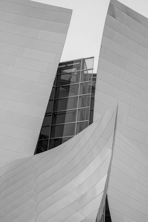 An intricate architectural structure is composed of interlocking metal panels and clear glass elements, forming fluid, abstract shapes. The design appears modern and futuristic, with smooth, reflective surfaces adding to its aesthetic.