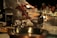 A bustling kitchen scene with chefs in white uniforms preparing food on a stainless steel countertop. Various cooking utensils, containers, and ingredients are spread across the surface, with an oil can prominently displayed. In the background, blurred figures of people suggest a lively dining atmosphere.