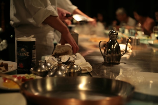 A bustling kitchen scene with chefs in white uniforms preparing food on a stainless steel countertop. Various cooking utensils, containers, and ingredients are spread across the surface, with an oil can prominently displayed. In the background, blurred figures of people suggest a lively dining atmosphere.