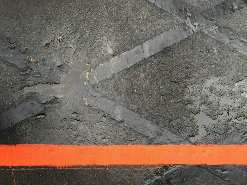 A rugged, textured surface with a bold red stripe running horizontally across it. The background features a dark, wet asphalt with intersecting faded lines, creating an industrial and urban feel.