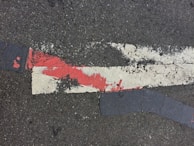 Rough asphalt surface with weathered, peeling paint, featuring a prominent red and white stripe crossing at an angle. The paint shows signs of wear and cracking, adding texture to the otherwise dark, gray ground.