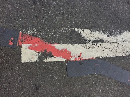 Rough asphalt surface with weathered, peeling paint, featuring a prominent red and white stripe crossing at an angle. The paint shows signs of wear and cracking, adding texture to the otherwise dark, gray ground.