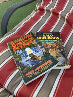 Two books titled 'Brown Girl in the Ring' by Nalo Hopkinson are placed on a fabric surface with red, beige, and white stripes. The covers of the books feature colorful and detailed illustrations.
