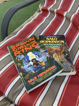Two books titled 'Brown Girl in the Ring' by Nalo Hopkinson are placed on a fabric surface with red, beige, and white stripes. The covers of the books feature colorful and detailed illustrations.