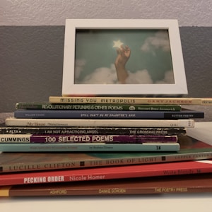 A stack of poetry books sits on a surface, with titles like 'Missing You, Metropolis' and 'Revolutionary Petunias & Other Poems.' On top of the stack is a framed photograph depicting a hand reaching up and holding a star against a cloudy sky.