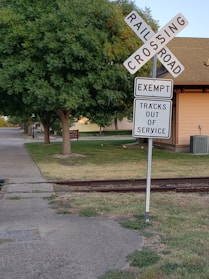 A quiet railroad crossing sign stands near an overgrown green area, with a sign indicating 'Tracks Out of Service.' A concrete path leads past the crossing. In the background, there's a residential house with a beige and brown exterior and a well-maintained lawn.
