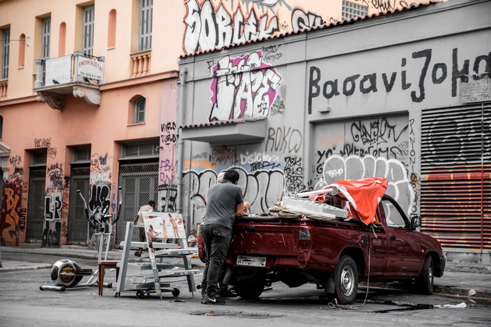 man in black jacket sitting on white wooden chair beside red car during daytime