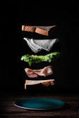 A deconstructed sandwich with its components floating above a wooden surface. The layers include two slices of brown bread, a piece of cheese, a layer of lettuce, and a slice of deli meat. Each ingredient is artfully suspended in mid-air, creating an impression of assembly in progress.