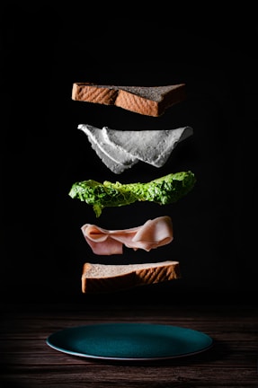 A deconstructed sandwich with its components floating above a wooden surface. The layers include two slices of brown bread, a piece of cheese, a layer of lettuce, and a slice of deli meat. Each ingredient is artfully suspended in mid-air, creating an impression of assembly in progress.
