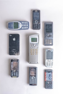 A collection of vintage mobile phones arranged neatly on a light-colored surface. The assortment includes various models, some with physical keypads and some with touchscreen capabilities. The phones exhibit signs of use, such as scratches and wear. The brands visible include Nokia, Sony Ericsson, and Apple, with the Apple phone showing a cracked screen.