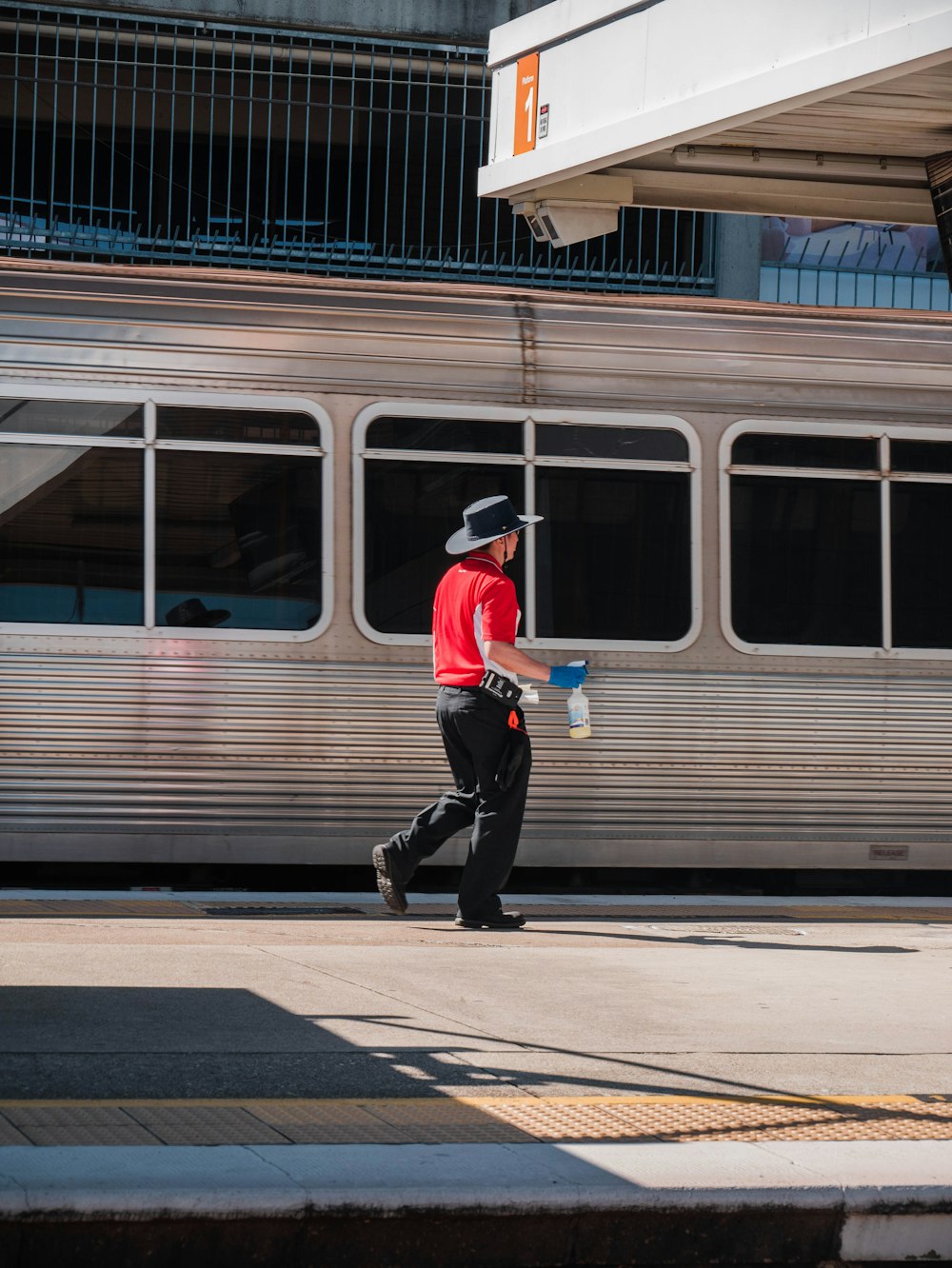 Hombre con camisa roja y pantalones negros caminando en la acera cerca de la estación de tren durante el día