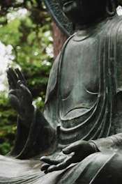 A bronze statue depicting a meditative figure with one hand raised and the other resting open on its lap. The background includes blurred greenery, indicating an outdoor setting.