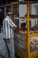 An older man is standing at a small food stall with a yellow frame, which is filled with stacks of round, bagel-like bread. He is wearing a striped shirt and jeans. The stall features various advertisements and graffiti. The setting appears to be an urban outdoor location with pavement and scaffolding visible.